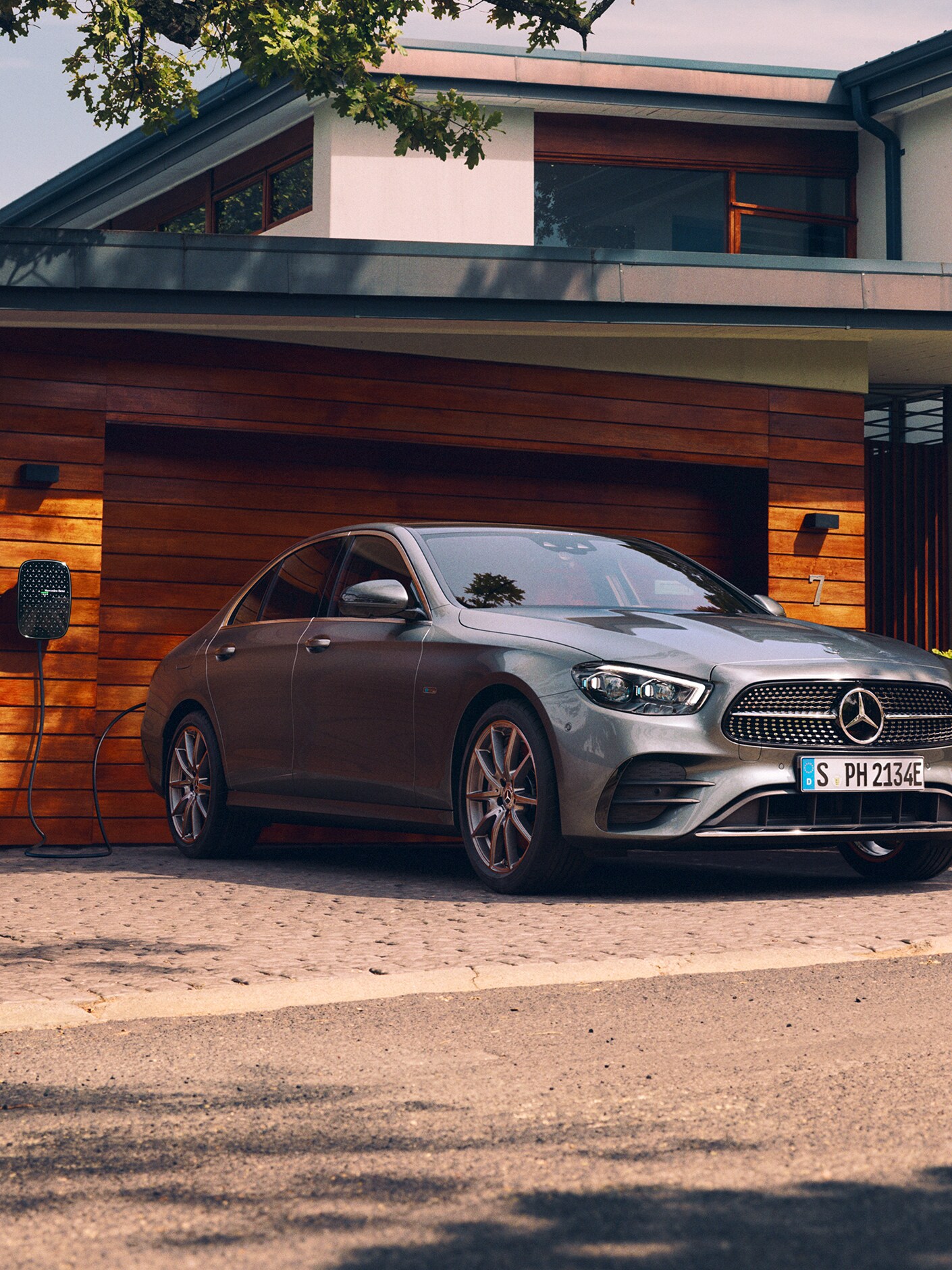 The image shows the Mercedes-Benz E-Class Saloon being charged outside a garage.