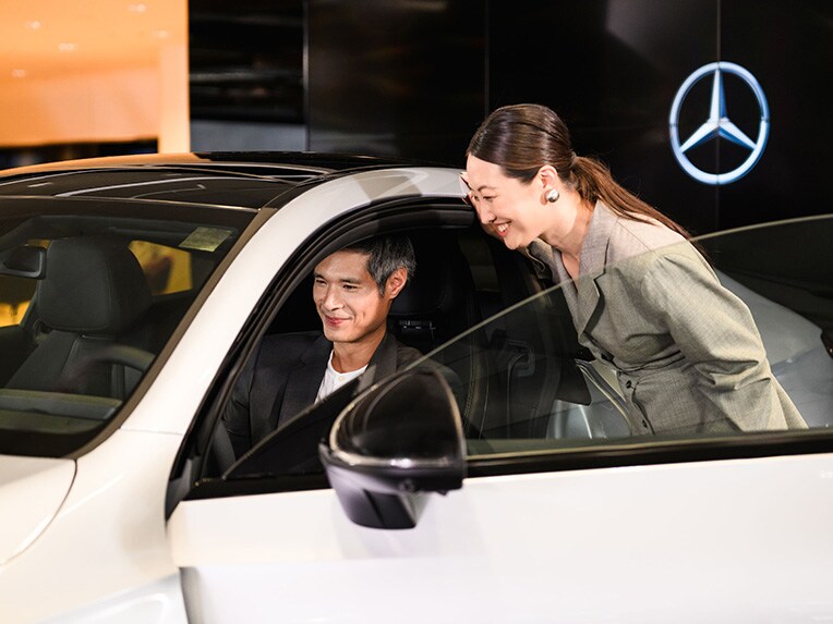 A person sitting in the drivers seat of a Mercedes-Benz vehicle with someone looking inside the passenger side door open.